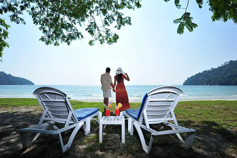 A couple enjoying a peaceful beach moment on Pangkor Island, with lounge chairs and drinks set under the shade, overlooking the calm sea.