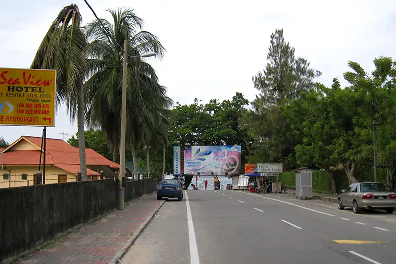 Street view leading to Pasir Bogak Beach on Pangkor Island with signage, palm trees, and hotels nearby.