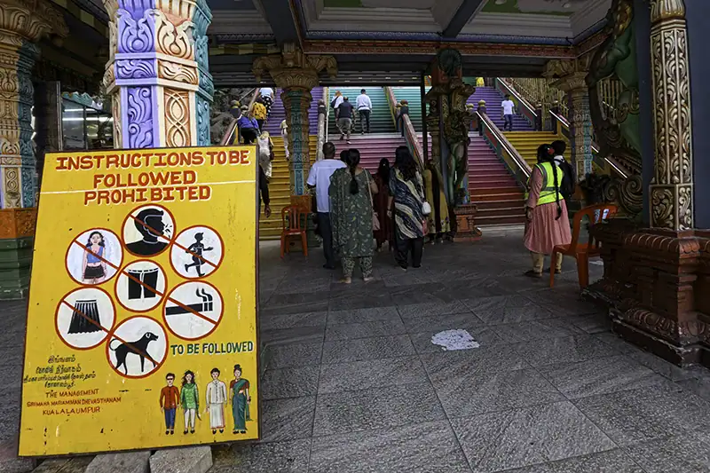 Signboard showing dress code instructions at Batu Caves with temple volunteers monitoring visitors near the staircase entrance.