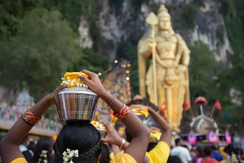 Devotees carrying milk pots during Thaipusam at Batu Caves in the evening, with Lord Murugan statue and colorful stairs in the background.