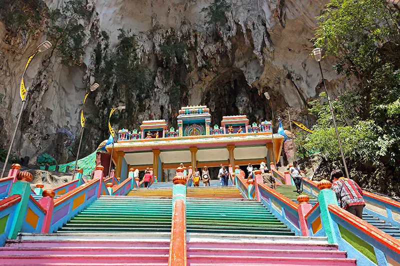Visitors reaching the top of the Batu Caves steps, standing near the colorful entrance to the Temple Cave, surrounded by towering limestone cliffs.