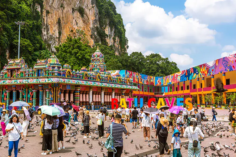 Tourists using umbrellas under the bright midday sun at Batu Caves, showing peak visiting hours when the temple grounds are busiest and hottest.